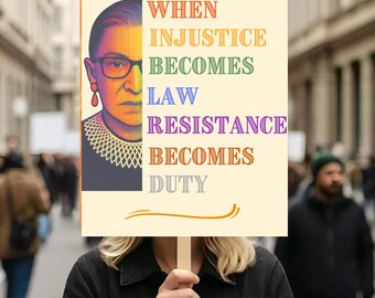 May include: A sign held by a person with the text "WHEN INJUSTICE BECOMES LAW RESISTANCE BECOMES DUTY". The sign features a colorful portrait of a woman with glasses and a black collar.
