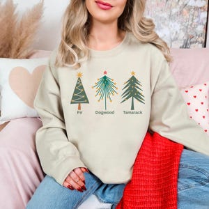 Può includere: Felpa beige con tre alberi di Natale stilizzati: Fir, Dogwood e Tamarack. Ogni albero ha una stella e dettagli decorativi. Le parole "Fir", "Dogwood" e "Tamarack" sono stampate sotto.