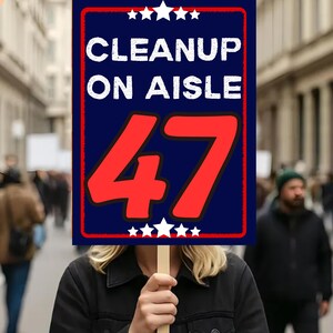 May include: A person holding a sign with the words "CLEANUP ON AISLE 47" in white and red text on a blue background. The sign has white stars at the top and bottom. The person is standing in a crowd.