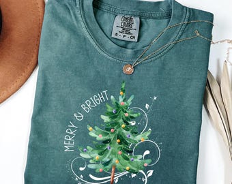 May include: A teal Comfort Colors t-shirt with a watercolor Christmas tree graphic. The tree is decorated with colorful lights and the words "Merry & Bright" are printed on the shirt. A gold necklace is draped across the shirt.