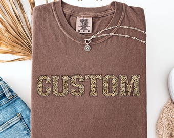 May include: A brown t-shirt with the word "CUSTOM" in leopard print letters. The shirt is made of a soft, comfortable material and has a relaxed fit.