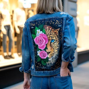 May include: Blue denim jacket with a hand-painted design on the back. The artwork features a leopard's face with blue eyes, pink roses, and white flowers. The jacket has a classic fit with pockets and button closures.