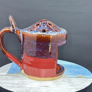 May include: A ceramic berry bowl with a handle, featuring a glossy glaze that transitions from a deep red at the base to a mottled purple and blue at the top. The bowl has a domed lid with several circular drainage holes and a small side handle.