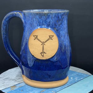 May include: A blue ceramic mug with a brown circle on the front. The circle has a design of three swords crossing in the center.