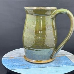 May include: Handmade ceramic mug with a large handle. The mug features a blend of green, blue, and brown hues. The base is a light brown color. The mug is sitting on a blue and white wooden surface.