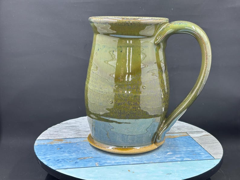 May include: Handmade ceramic mug with a large handle. The mug features a blend of green, blue, and brown hues. The base is a light brown color. The mug is sitting on a blue and white wooden surface.