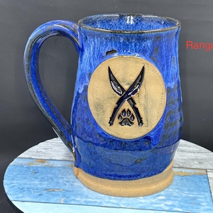 May include: A handcrafted blue ceramic mug with a large handle. The mug features a tan emblem with crossed knives and a paw print. The mug has a blue glaze with darker blue and black accents. The base of the mug is tan.