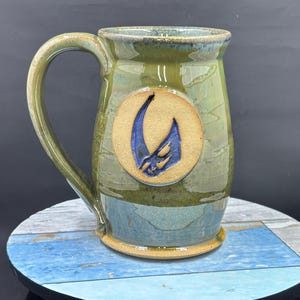 May include: Handmade ceramic mug with a large handle. The mug features a green and blue glaze with a circular, tan-colored emblem. The emblem has a blue, abstract design. The mug is sitting on a round, multi-colored surface.