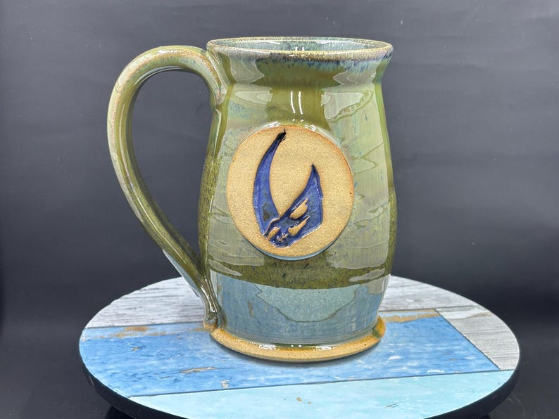 May include: Handmade ceramic mug with a large handle. The mug features a green and blue glaze with a circular, tan-colored emblem. The emblem has a blue, abstract design. The mug is sitting on a round, multi-colored surface.