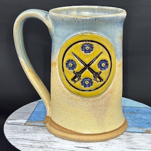 May include: A handmade ceramic mug with a large handle. The mug features a yellow emblem with crossed swords and blue floral accents. The mug has a light blue and yellow glaze.