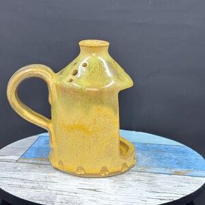 May include: A yellow ceramic oil burner with a glossy finish, featuring a curved handle on the side, a small chimney-like top with three ventilation holes, and a wide arched opening at the base for placing a tea light candle inside.