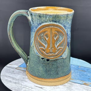 May include: A large ceramic mug with a handle, featuring a blue and green glaze with a brown emblem. The mug has a wide base and a textured surface, perfect for enjoying a beverage. The mug is approximately 6 inches tall.