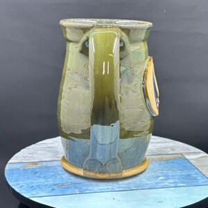 May include: A handmade ceramic mug with a large handle. The mug features a blend of green, blue, and brown glaze. A small, decorative element is attached to the side. The mug sits on a round, multi-colored surface.