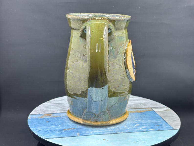 May include: A handmade ceramic mug with a large handle. The mug features a blend of green, blue, and brown glaze. A small, decorative element is attached to the side. The mug sits on a round, multi-colored surface.