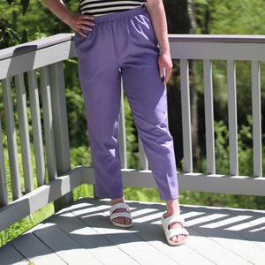 May include: A pair of lavender trousers with a wide waistband and a relaxed fit. The trousers are being worn by a person standing on a wooden deck.