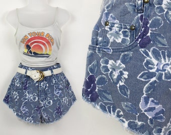 May include: A pair of blue denim shorts with a floral print. The shorts have a high waist and a frayed hem. The shorts are worn with a white tank top that says "Sun Your Buns" and a white belt.