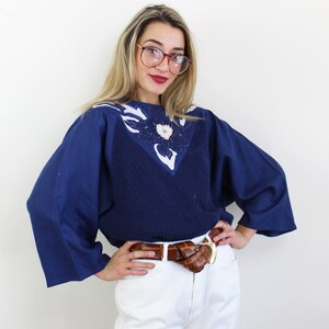 May include: A navy blue sweater with a white and blue floral design on the neckline. The sweater has a relaxed fit and long sleeves. The model is wearing a brown leather belt with a gold buckle.