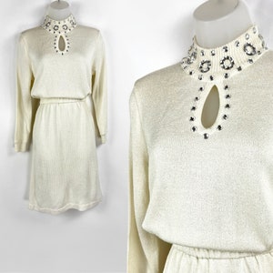 May include: A white knit dress with a high mock neck and a keyhole cutout at the neckline. The neckline is embellished with a row of silver rhinestones.