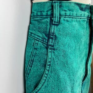 May include: Close-up of teal acid wash denim jeans with a classic five-pocket design. The jeans have a high waist and a button and zipper closure. The top of the jeans are paired with a black and cream striped top.