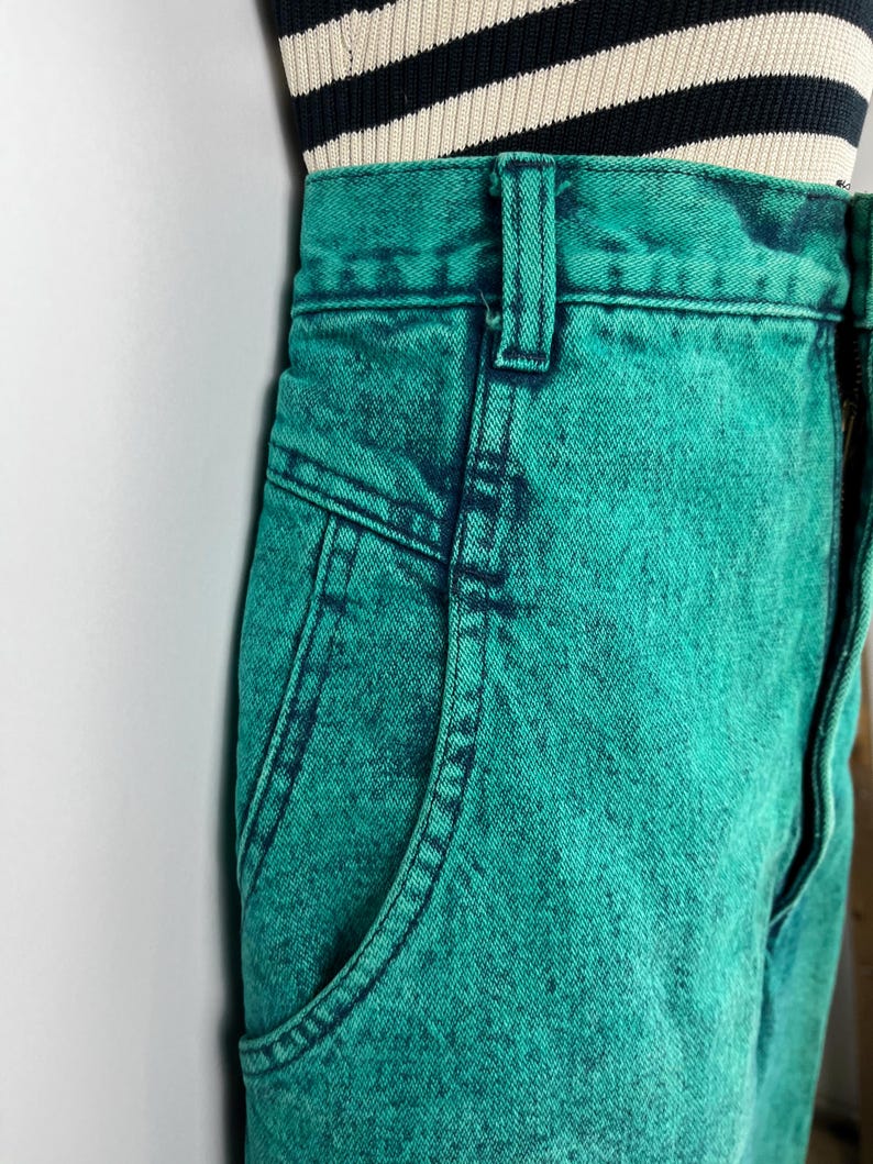 May include: Close-up of teal acid wash denim jeans with a classic five-pocket design. The jeans have a high waist and a button and zipper closure. The top of the jeans are paired with a black and cream striped top.