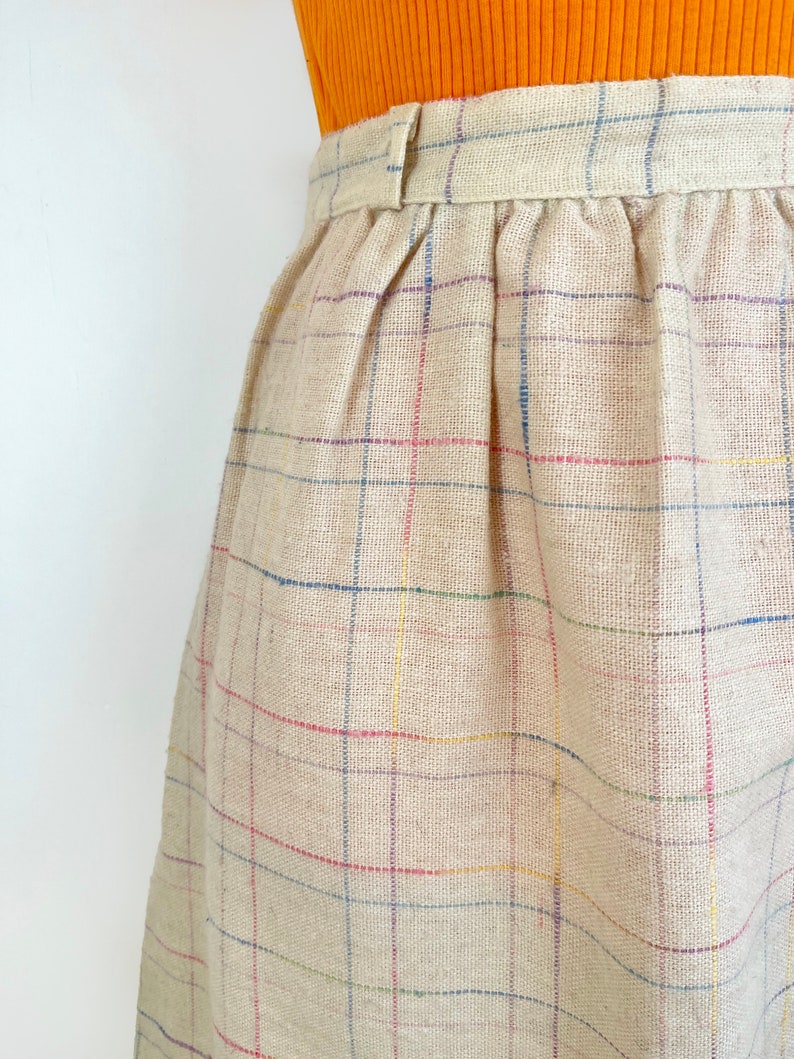 May include: A beige skirt with a multi-colored plaid pattern. The skirt has a waistband with a button closure.