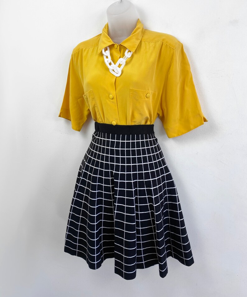 May include: A yellow short-sleeved button-down shirt with a white chain necklace and a black and white checkered skirt.