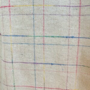 May include: A close-up of a beige fabric with a woven texture. The fabric has a checkered pattern with thin lines of red, yellow, blue, green, and purple.