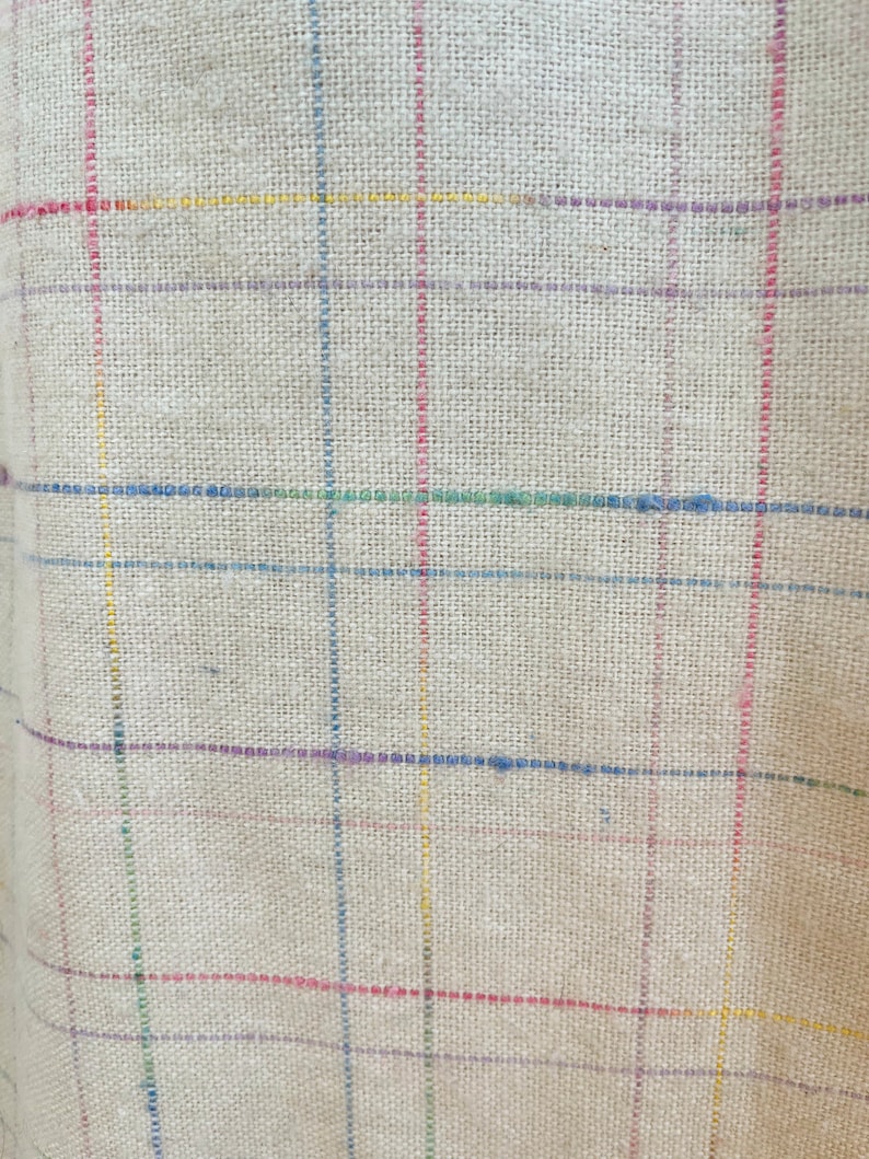 May include: A close-up of a beige fabric with a woven texture. The fabric has a checkered pattern with thin lines of red, yellow, blue, green, and purple.