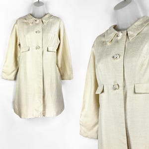 May include: A cream-colored, long-sleeved coat with a collared neckline and button closures. The coat has two pockets on the front and a slightly flared hem.