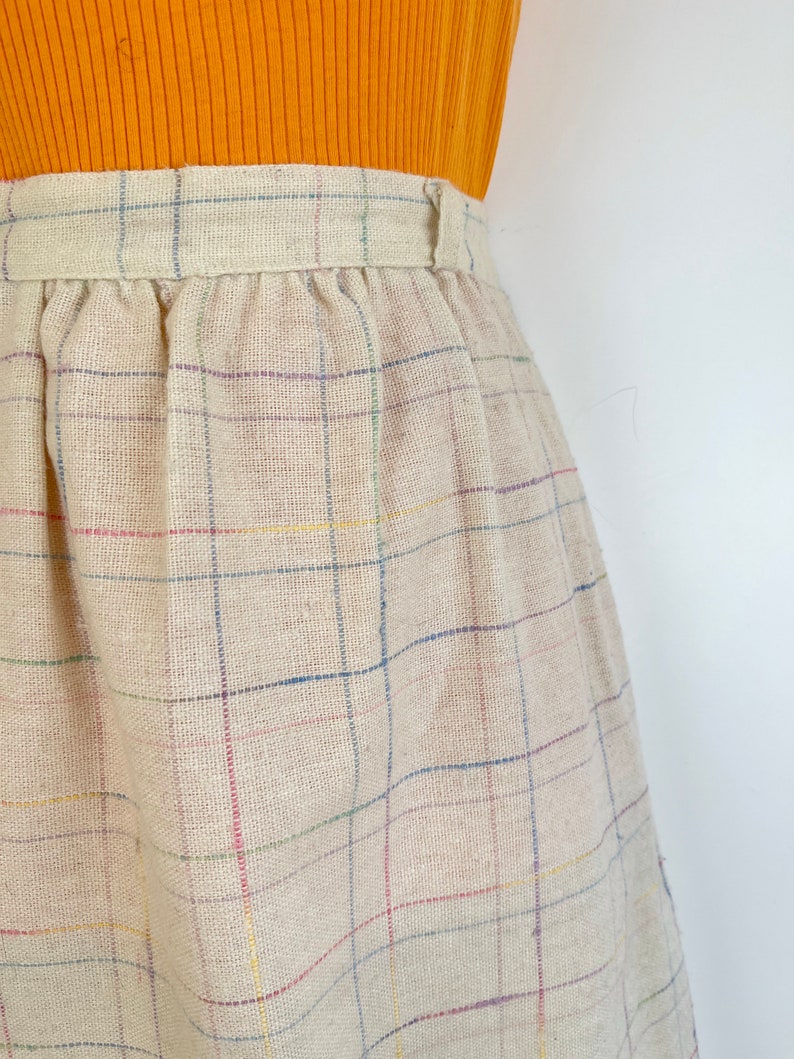May include: A beige skirt with a multi-colored plaid pattern. The skirt has a waistband with a button closure.