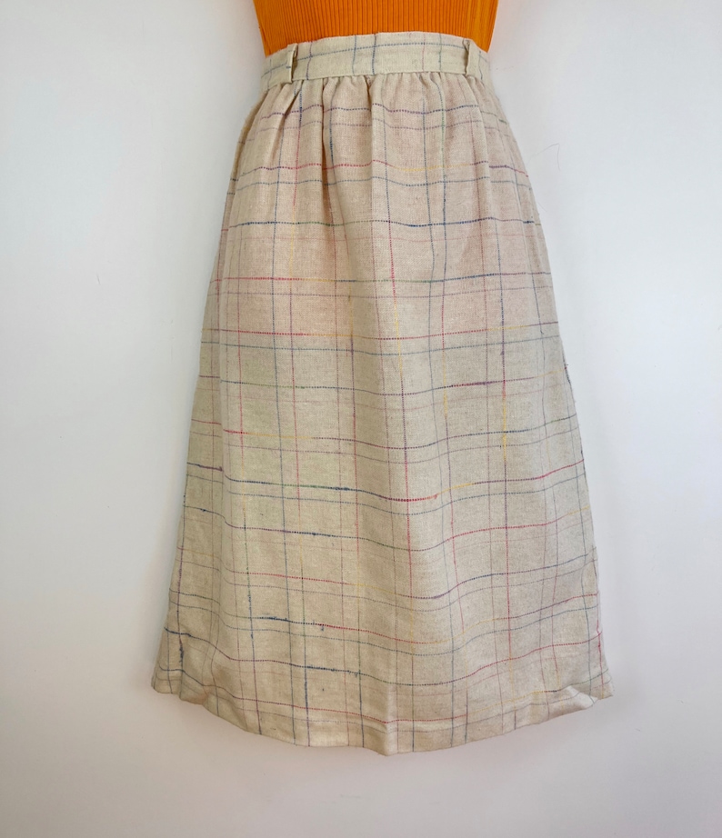 May include: A beige midi skirt with a plaid pattern in shades of blue, red, and yellow. The skirt has a high waist and a straight silhouette.
