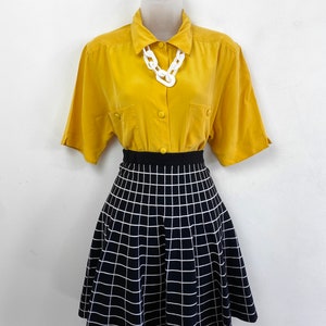 May include: A yellow button-down shirt with short sleeves and a white plastic chain necklace. The shirt is paired with a black and white checkered skirt.