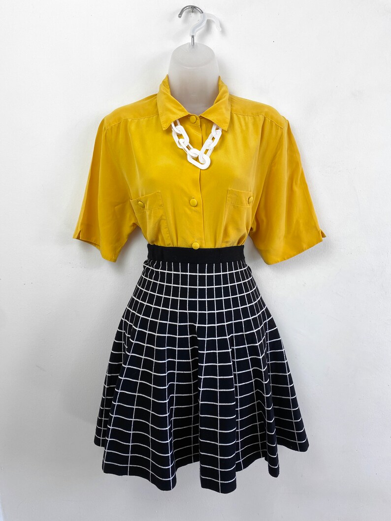 May include: A yellow button-down shirt with short sleeves and a white plastic chain necklace. The shirt is paired with a black and white checkered skirt.