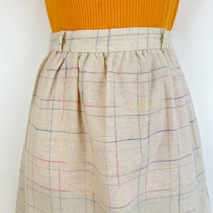 May include: A beige plaid skirt with thin multi-colored stripes. The skirt has a waistband with a button closure.