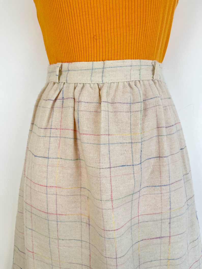 May include: A beige plaid skirt with thin multi-colored stripes. The skirt has a waistband with a button closure.