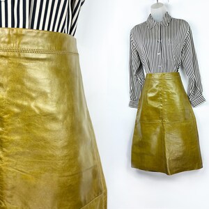 May include: A-line skirt in a golden-olive hue. The skirt is made of a smooth, possibly leather, material. The skirt is paired with a black and white striped button-down shirt. The skirt has a classic, vintage aesthetic.