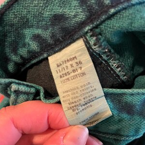 May include: Close-up of a pair of green and black tie-dye denim jeans. The jeans have a tag that reads "KA7260M 11/12 X 36 A265-017 100% COTTON". The tag also includes washing instructions. The jeans are made in the USA.