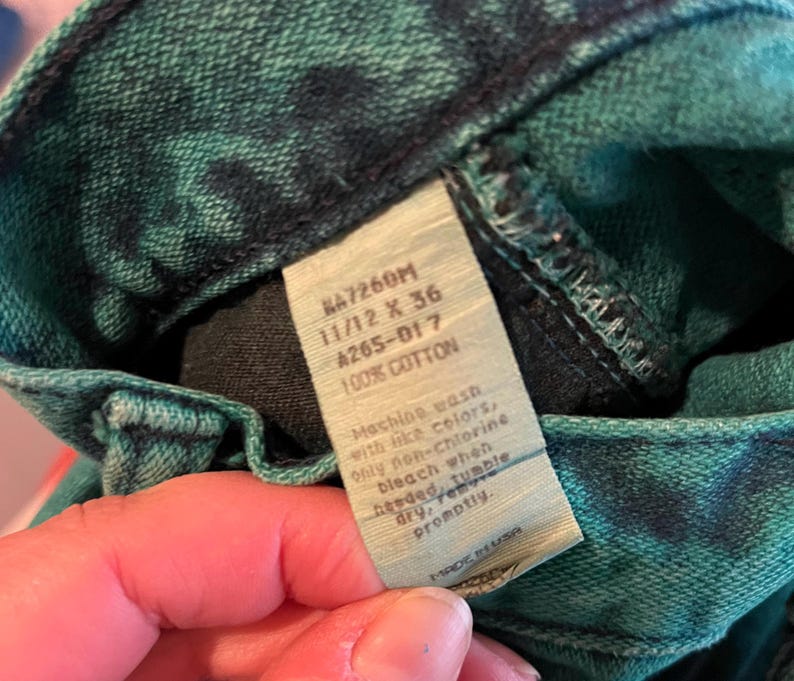 May include: Close-up of a pair of green and black tie-dye denim jeans. The jeans have a tag that reads "KA7260M 11/12 X 36 A265-017 100% COTTON". The tag also includes washing instructions. The jeans are made in the USA.