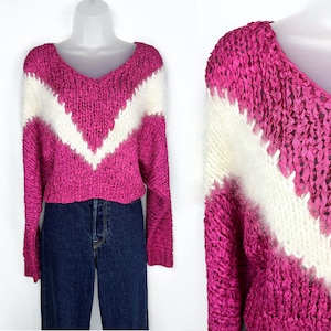 May include: A pink and white knit sweater with a V-shaped design. The sweater is made of a chunky yarn and has a loose fit.