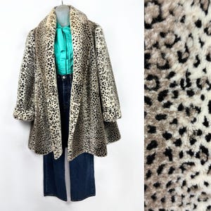 May include: A vintage leopard print coat with a wide collar and long sleeves. The coat is displayed on a mannequin with a teal blouse and dark wash jeans. The coat's pattern features black spots on a beige and cream background.