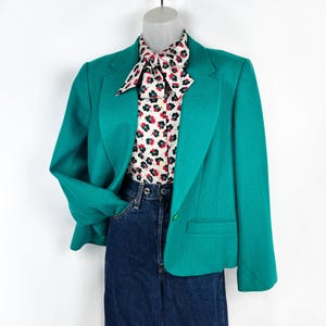 May include: A teal blazer with a button closure, paired with a floral print blouse and dark wash denim jeans. The blouse features a white background with red and black floral patterns. The blazer has a classic lapel and a tailored fit.