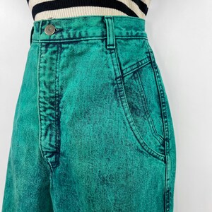 May include: Close-up of high-waisted, acid-wash teal jeans. The jeans have a button closure, belt loops, and front pockets. The fabric has a textured appearance. The top of a black and white striped shirt is visible.