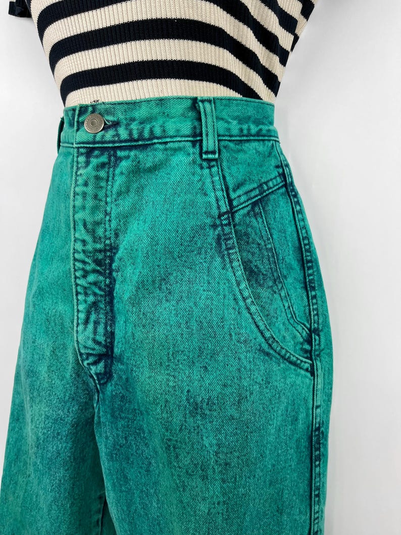 May include: Close-up of high-waisted, acid-wash teal jeans. The jeans have a button closure, belt loops, and front pockets. The fabric has a textured appearance. The top of a black and white striped shirt is visible.