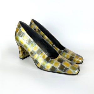 May include: A pair of square-toed pumps with a checkerboard pattern in shades of olive green, gold, and brown. The heels are chunky and the shoes have a glossy finish. The shoes are set against a white background.