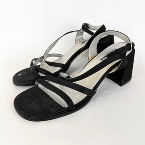 May include: Black heeled sandals with multiple straps and a buckle closure. The sandals have a block heel and are made of a suede-like material.