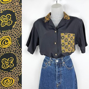 May include: Black short-sleeved button-down shirt with a patterned collar and pocket. The collar and pocket feature a repeating yellow and brown circular design. The shirt is paired with blue denim jeans.