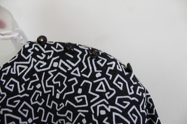 Puede incluir: Tela negra y blanca con un patr&oacute;n geom&eacute;trico. La tela tiene una superficie texturizada y est&aacute; decorada con peque&ntilde;os botones negros.