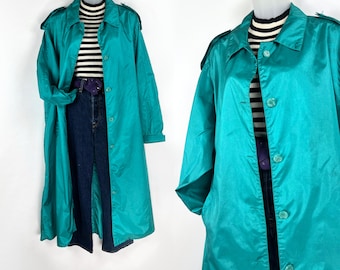May include: A teal-colored trench coat with a button-up front and a collared neckline. The coat is displayed on a mannequin, paired with a black and white striped top and dark blue jeans. The coat has a shiny, satin-like finish.