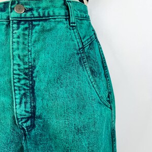 May include: Close-up of high-waisted, acid-wash teal denim jeans. The jeans have a button closure, belt loops, and pockets. The fabric has a textured appearance with darker teal accents. A black and white striped top is partially visible.