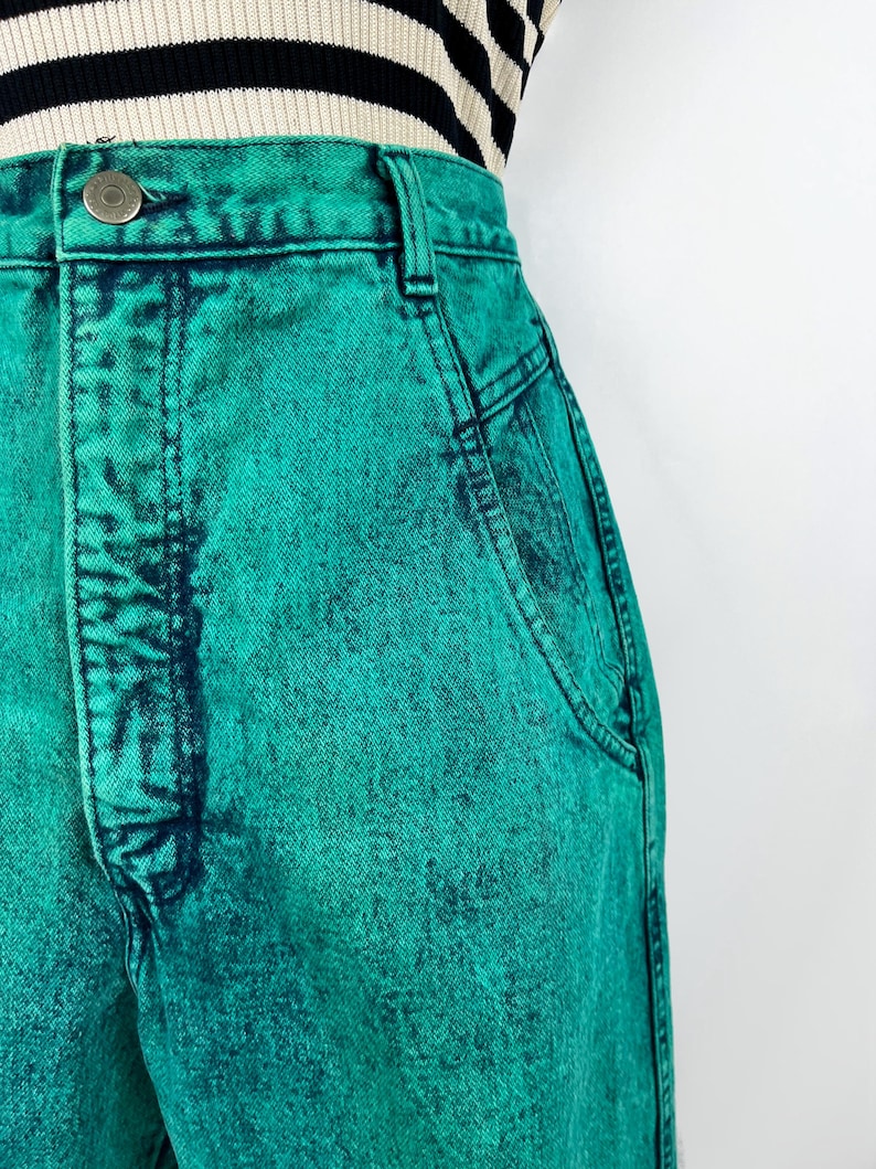 May include: Close-up of high-waisted, acid-wash teal denim jeans. The jeans have a button closure, belt loops, and pockets. The fabric has a textured appearance with darker teal accents. A black and white striped top is partially visible.